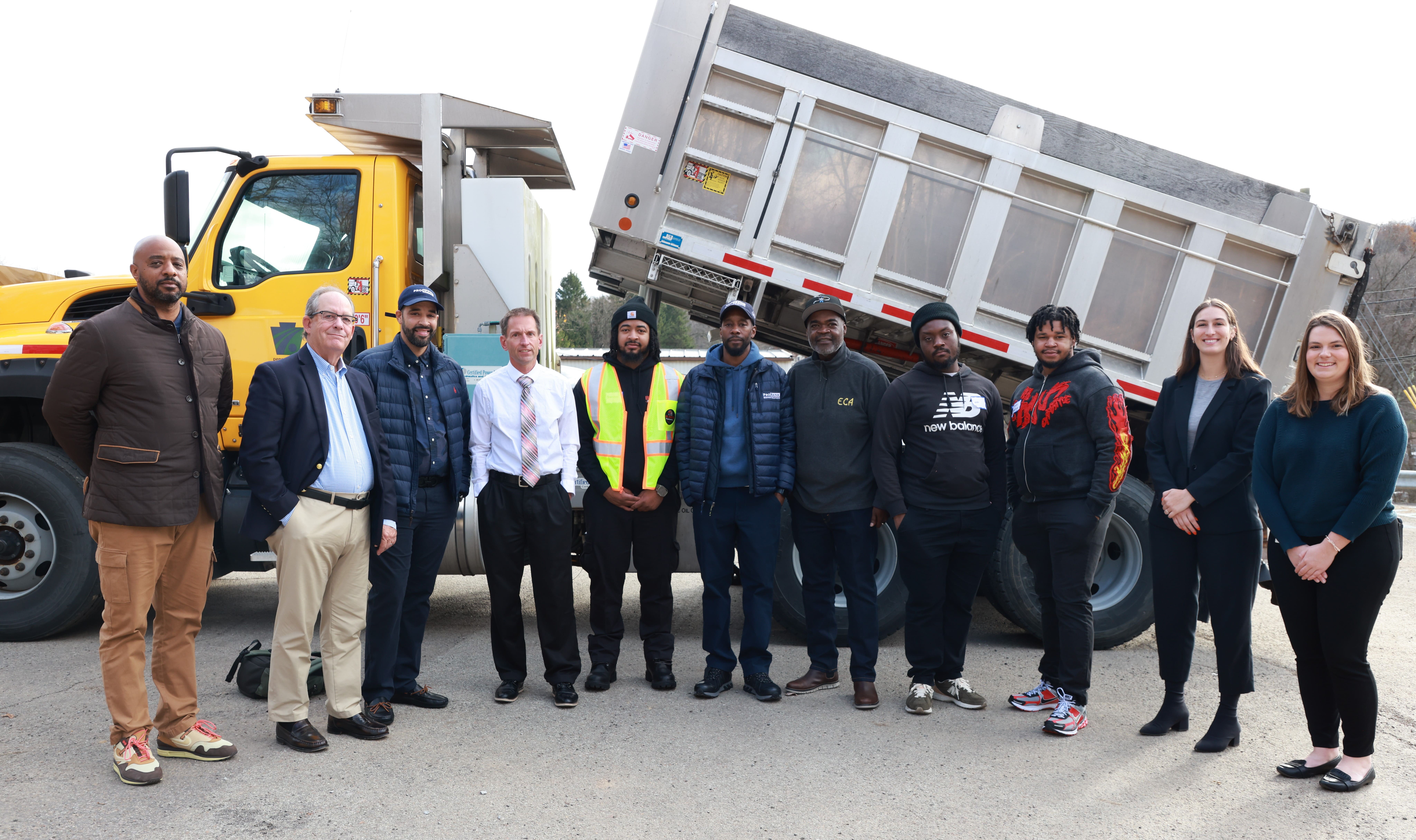 A group of 11 individuals stand in front of a yellow PennDOT dump truck in a parking lot.